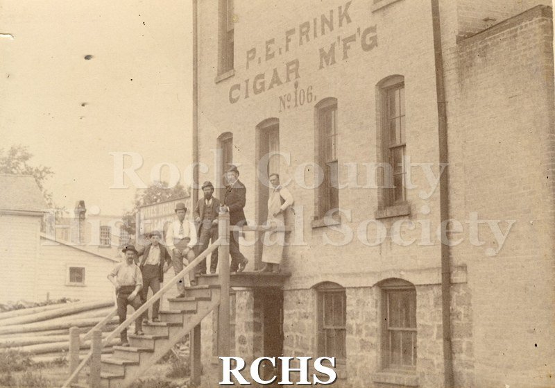Black and white photograph of six unidentified men standing on the steps of a cigar warehouse. The sign on the building reads: "P. E. Frink Cigar M'F'G No. 106".