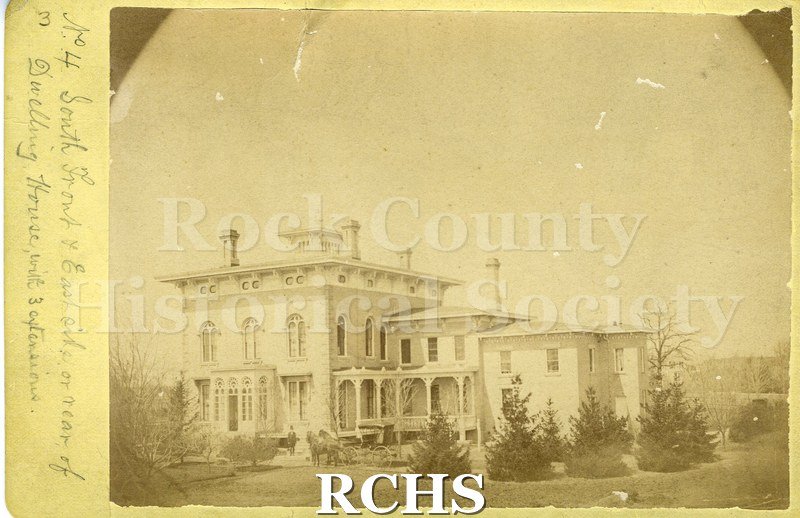 Black and white photograph of Lincoln Tallman House showing the front porch and conservatory sides. Shrubs along front of house, trees in front and back.