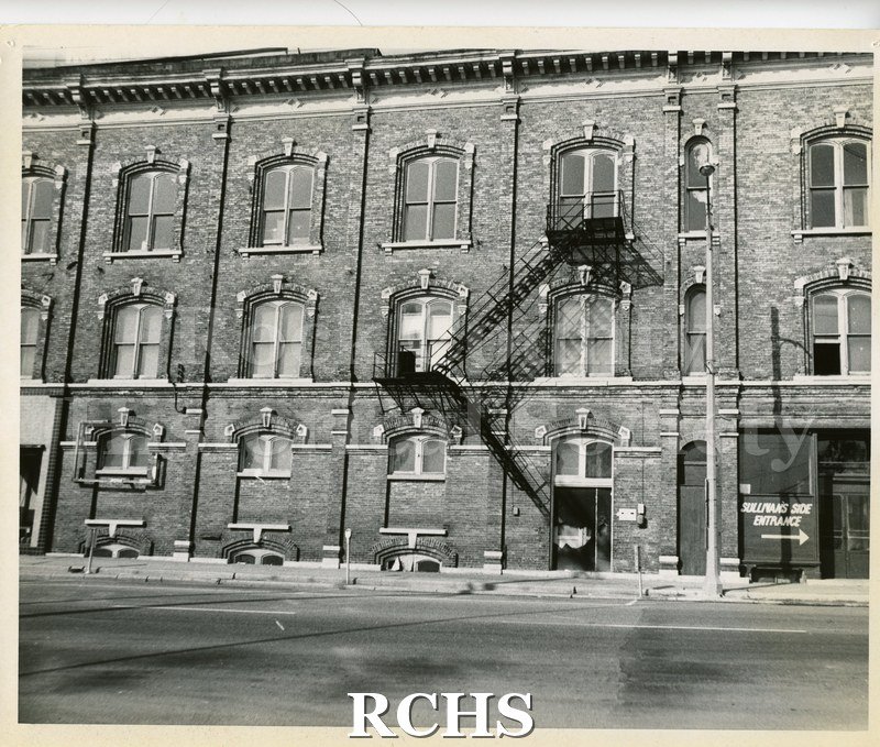 Black and white photograph of the back of a three story brick building with a metal fire excape on the side.