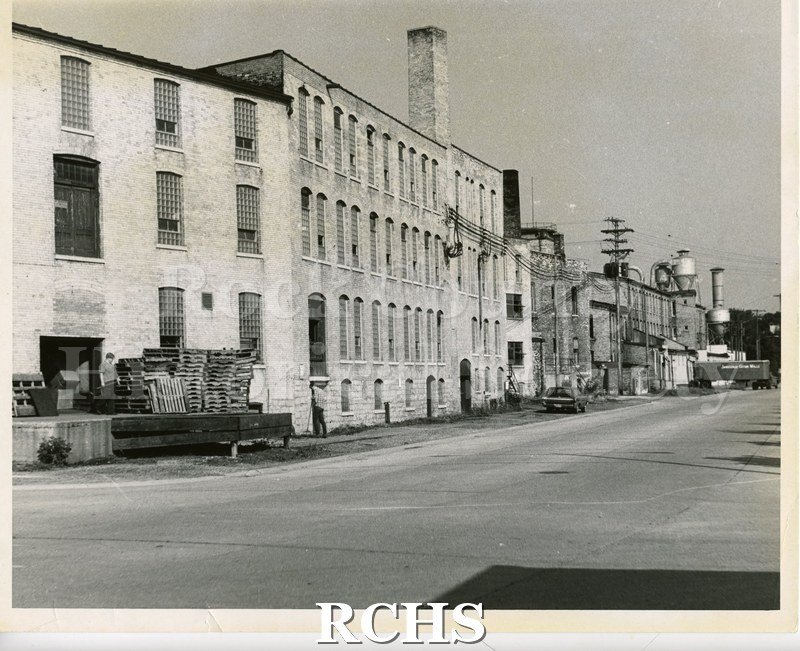 Black and white photograph of Rock River Manufacturing. Shows a four story factory building with a chimney; loading dock and railroad car with a sign: "Janesville.