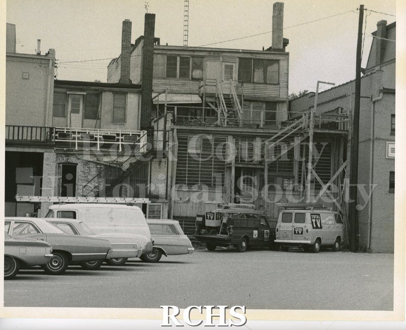 Black and white photograph of the backs of several building on Main Street. There are two trucks parked with "Total TV" on the sides.