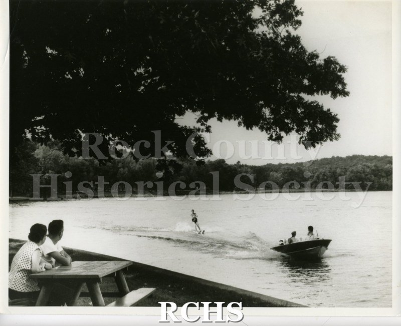 Black and white photograph of two people sitting at a picnic table watching a water skier being pulled by a boat with two people in it.