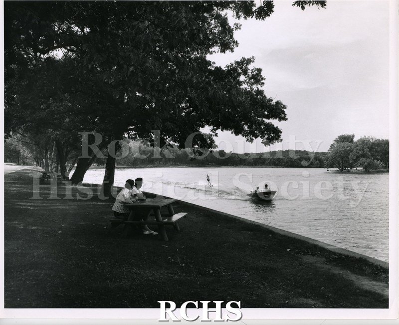 Black and white photograph of two people sitting at a picnic table watching a water skier being pulled by a boat with two people in it.