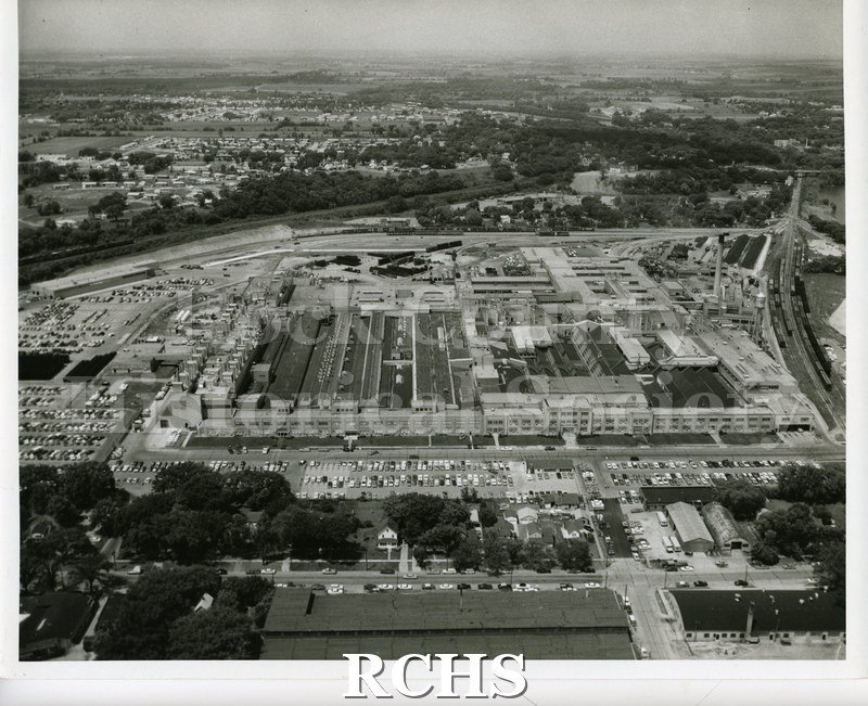 Black and white aerial photograph of the General Motors Plant. Shows buildings and cars parked as well as other buildings in the background.