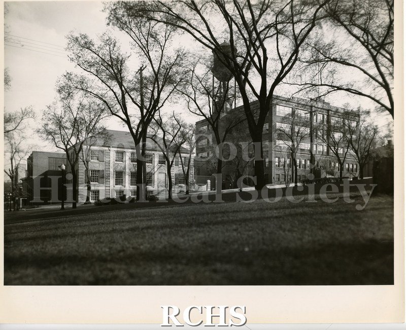 Black and white photograph of the Parker Pen plant. A four story building with a water tower above it. To the left is a three story limestone building.