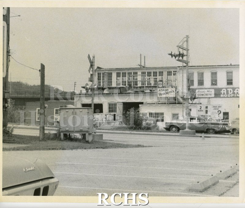 Black and white photograph of Adams Roofing, N. Main. Shows a two story building with signs: "Adams Ready-mix"; "Ready Mix Concrete; and "Adams Roofing".