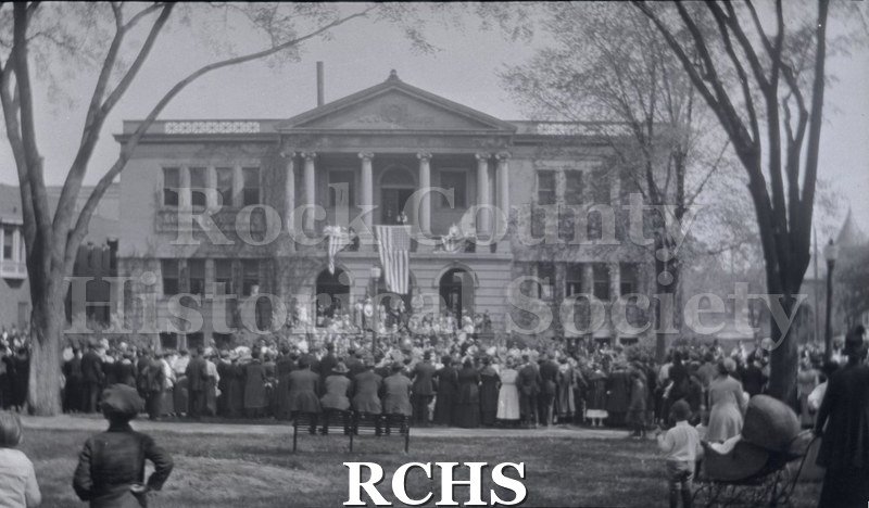Black and white photograph of a large crowd of people standing in front of a two story building with a balcony.