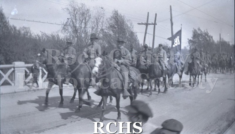Black and white photograph of a large crowd of a long line of horses and riders parading down a street.