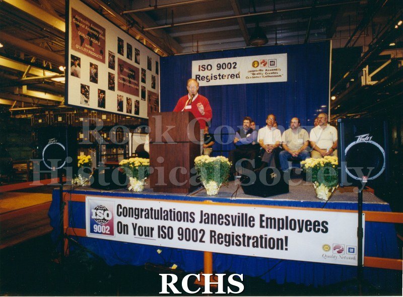 Historic image of Color: GM Unknown employees on stage/presenting banner says Congradulations Janesville employees on your 9002 Registration -1 man presenting in.
