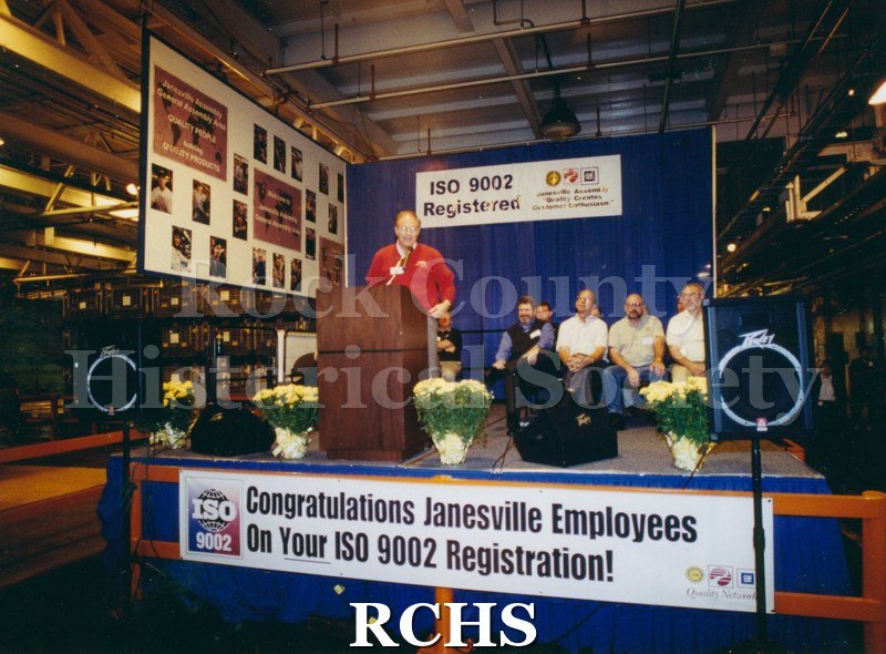 Historic image of Color: GM Unknown employees on stage/presenting banner says Congradulations Janesville employees on your 9002 Registration -1 man presenting in.