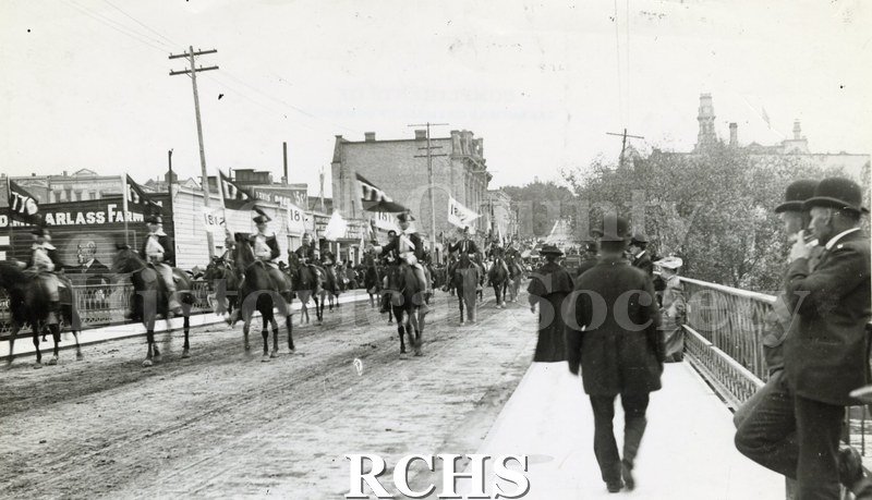 Historic image of Memorial Day Parade late 1800's. Shows men on horseback parading over Court Street bridge, Janesville, carrying flags reading "1776" and "1812".