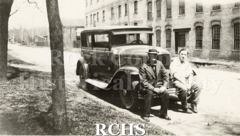 Historic image of Harry McKinney and Ithamar Sloan. Two men are seated on the front bumper of an old automobile. Three story brick building in background.