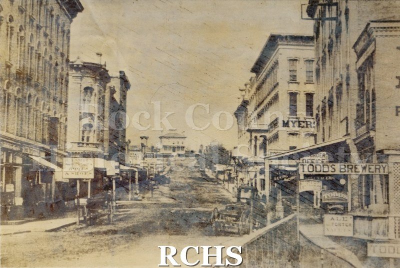 Grayscale. Image depicts a downtown. Businesses and vehicles can be seen on either side of the road.