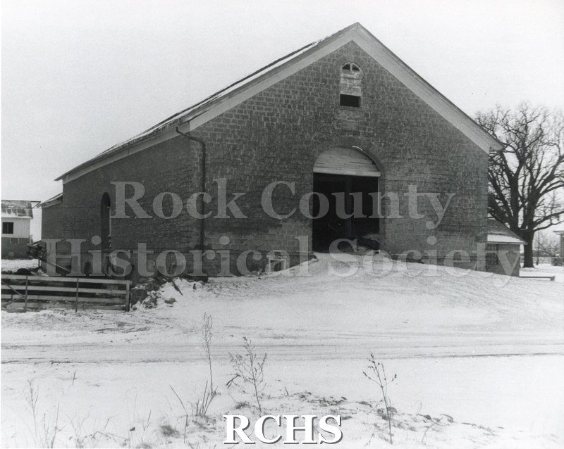 Black and white photograph of Wyman-Rye Farmstead Barn, a stone barn with an open door into the haymow. There is snow on the ground.