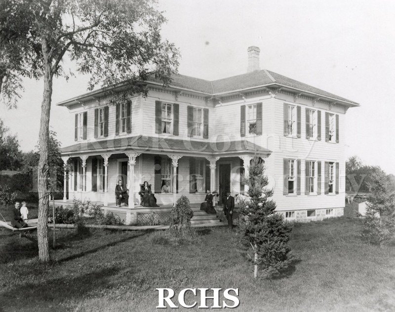 Black and white photograph of the Ole Nattestad house, a two story house with a pillared porch. Several people are pictured on the porch and yard.