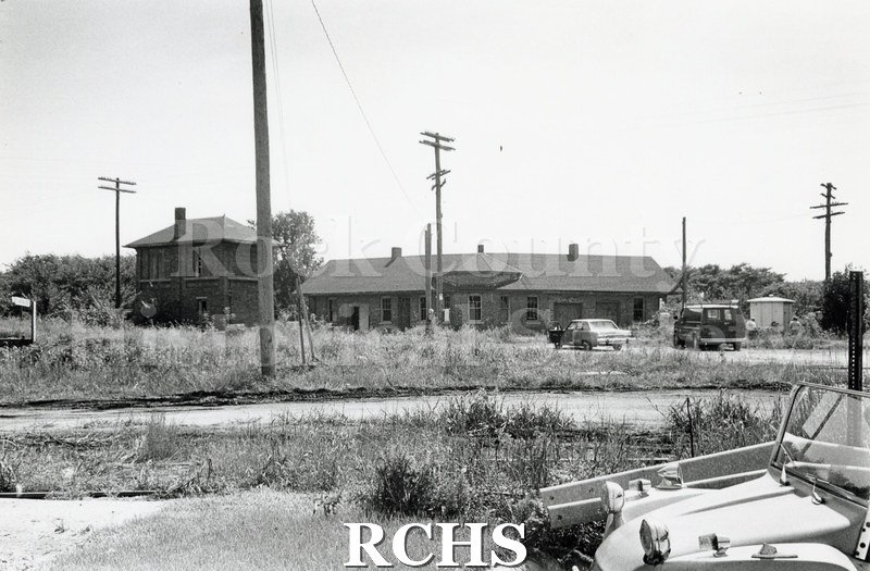 Black and white photograph of a two story small railroad building on the left and a one story depot on the right.