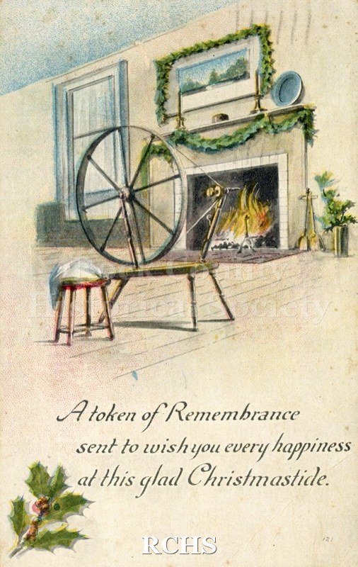 Color postcard of "old time" spinning wheel and stool in front of fire in fireplace with greenery draped around mantle.