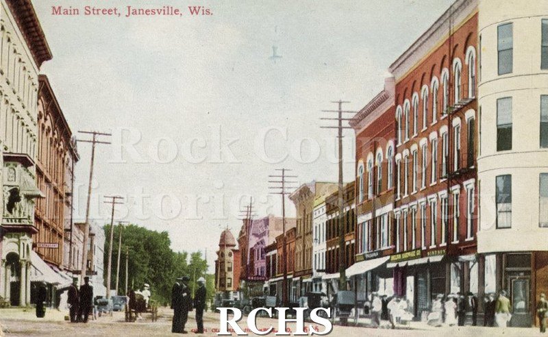 Postcard. Colored image of Main Street in Janesville. The image was taken on a busy day.