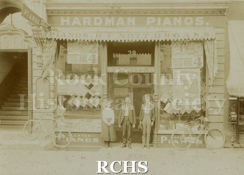 Tasie Nott, Henry F. Nott, and Chas. Kemp. sepia photo of a storefront with three people standing in the doorway. Above the doorway is a sign: "Hardman Pianos".